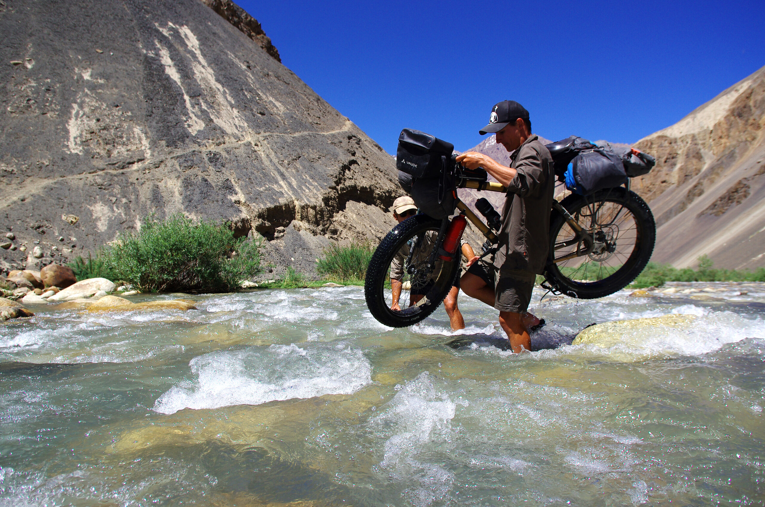 Brian Mathé, assisté de Morgan Monchaud, traverse la rivière en portant son vélo en bambou utilisé pour traverser le Pamir via des sentiers difficiles